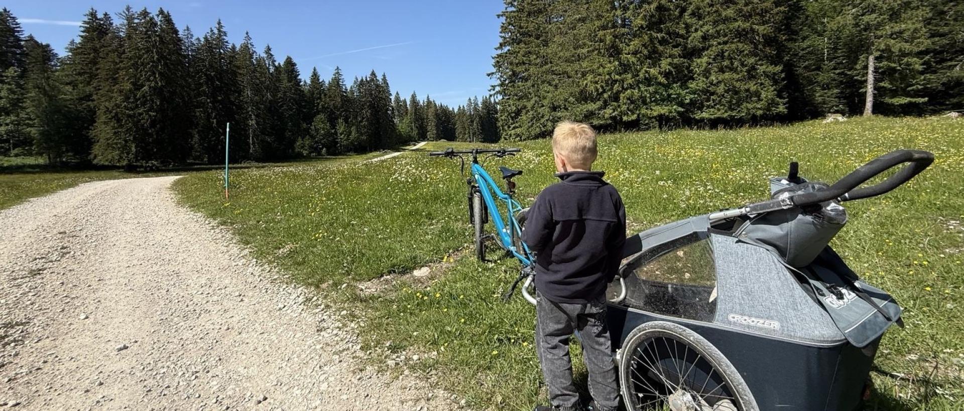 Enfant devant un vélo et une remorque sur les chemins forestiers du Jura suisse près des Cernets